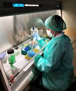 Siedząca kobieta w seledynowym kombinezonie i jasnej maseczce. Trzyma w dłoniach mały pojmenik i urządzenie przypominające strzykawkę. Widok fragmentu laboratorium. . Kliknij, aby powiększyć zdjęcie.